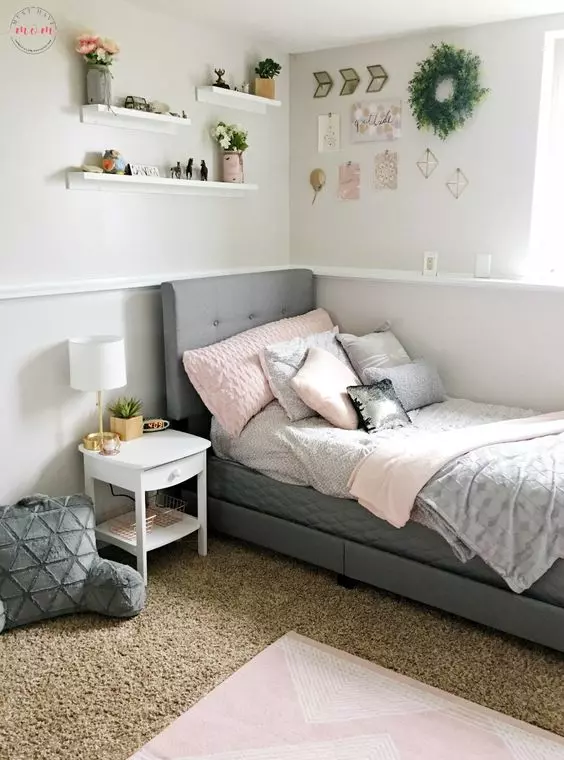 a modern teen bedroom with a grey upholstered bed and pastel bedding layered rugs wall mounted shelves and a greenery wreath