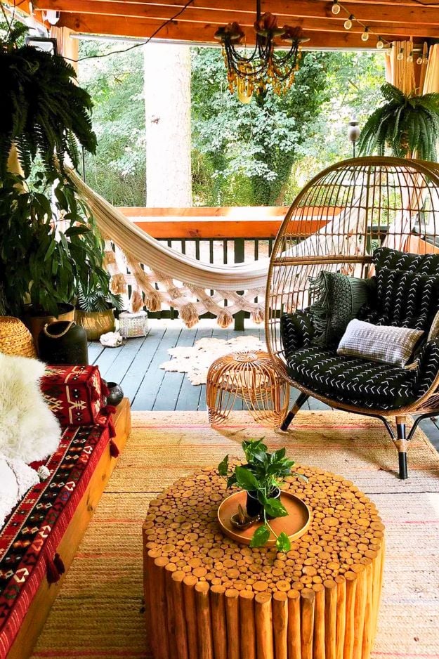 Cozy covered backyard deck with a wicker egg chair, hanging hammock, wooden coffee table, layered rugs, lush plants, and warm string lights creating a relaxed boho outdoor lounge.