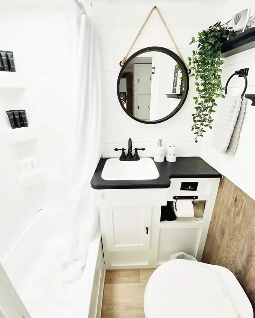 Small camper bathroom with a teal vanity cabinet, light wood countertop, white vessel sink, black faucet, round black-framed mirror, hexagon tile wall, woven storage basket, potted plant, white toilet, and simple modern decor in a bright, cozy space.
