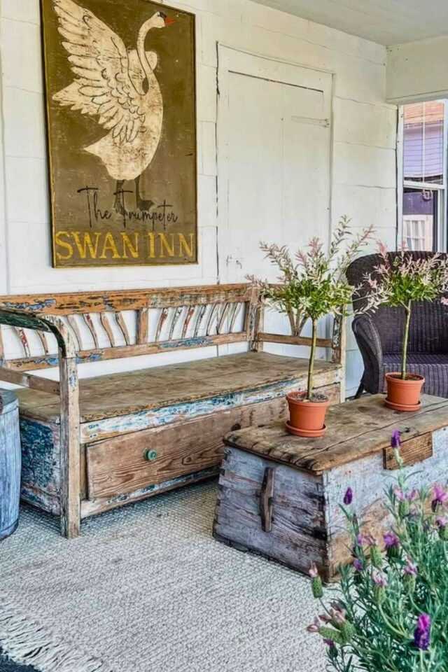 Rustic small front porch with a weathered wooden bench, an antique trunk used as a coffee table, potted plants on top, a wicker chair, and a vintage swan wall sign against white shiplap walls, creating a cozy, charming farmhouse feel.
