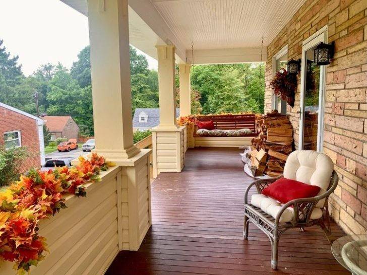 Cozy small front porch with white columns, fall leaf garland along the railing, a wooden porch swing with cushions, wicker chair, and stacked firewood against a brick wall.