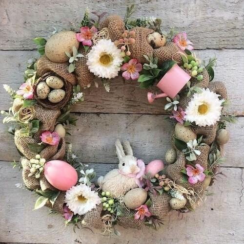 Rustic Easter wreath made from burlap wrapped around a grapevine base, decorated with pastel eggs, pink flowers, greenery, and a small bunny figure at the bottom, hanging on a weathered wood background.