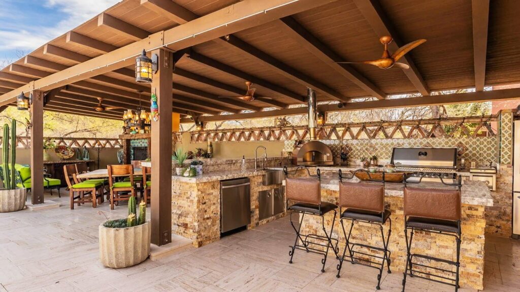 Covered outdoor Mexican kitchen with Spanish hacienda style, featuring a stone bar with metal stools, built-in grill and wood-fired oven, patterned tile backsplash, wooden pergola ceiling with fans, potted cacti, and a cozy dining area in a sunlit courtyard.