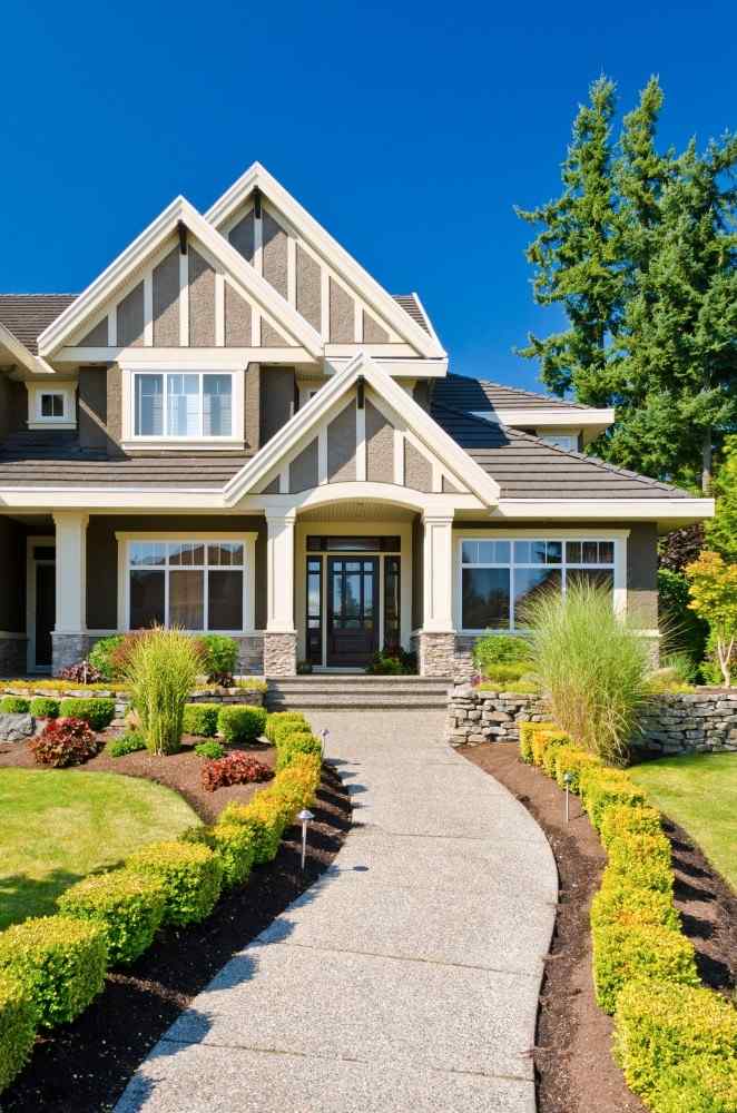 A charming Craftsman-style home with a curved concrete walkway, neatly trimmed hedges, layered garden beds, and lush green landscaping under a bright blue sky.