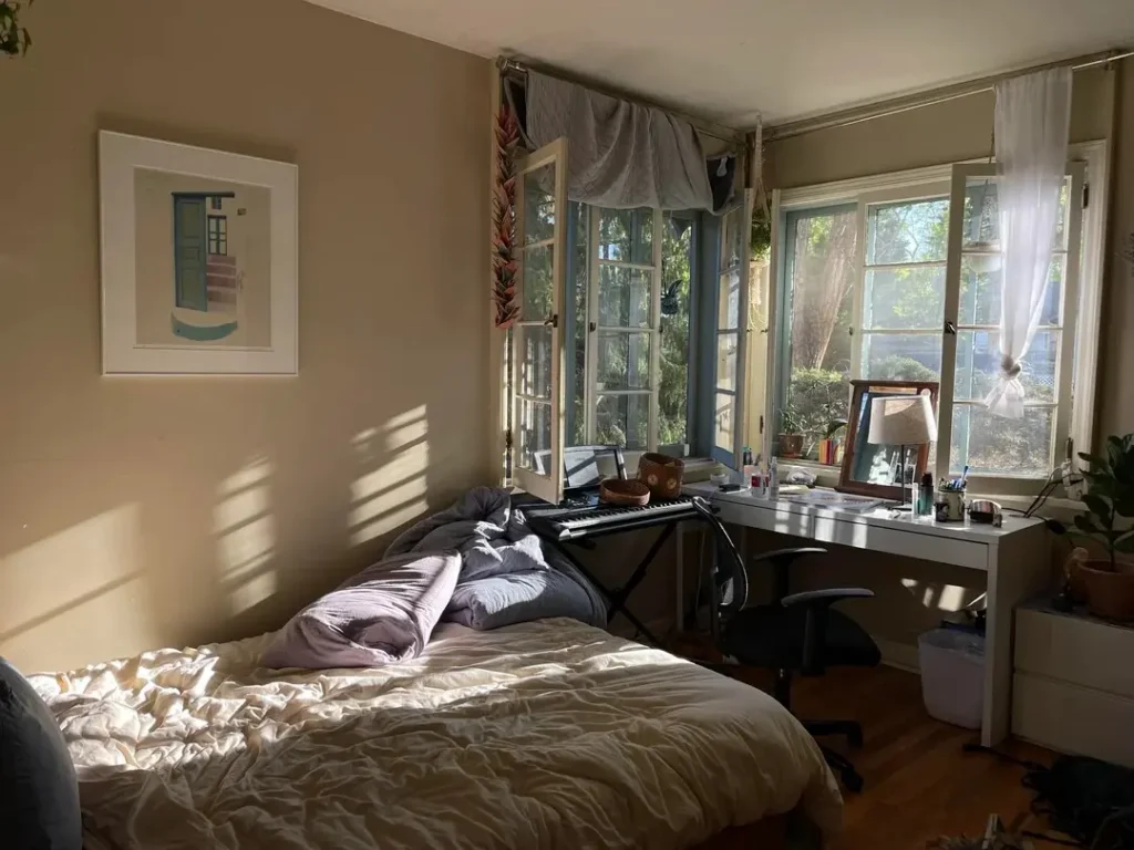 Sunlit bedroom with beige walls, an unmade bed in neutral bedding, and tall French windows casting afternoon light across the room. A white desk with a mirror, plants, and art supplies sits by the windows, creating a cozy, creative workspace.