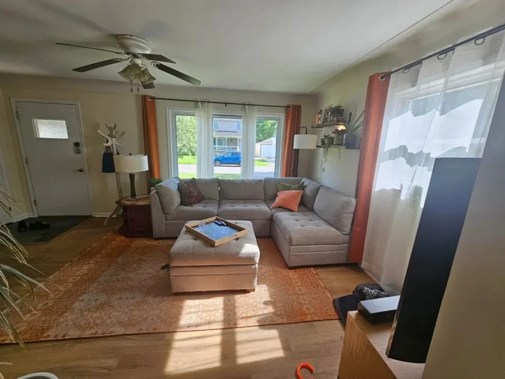 Bright living room with a gray L-shaped sectional sofa and matching ottoman on a patterned orange rug. Large windows with sheer white curtains and rust-colored drapes let in natural light. A ceiling fan hangs overhead, with a side table, lamps, plants on wall shelves, and a TV partially visible on the right.
