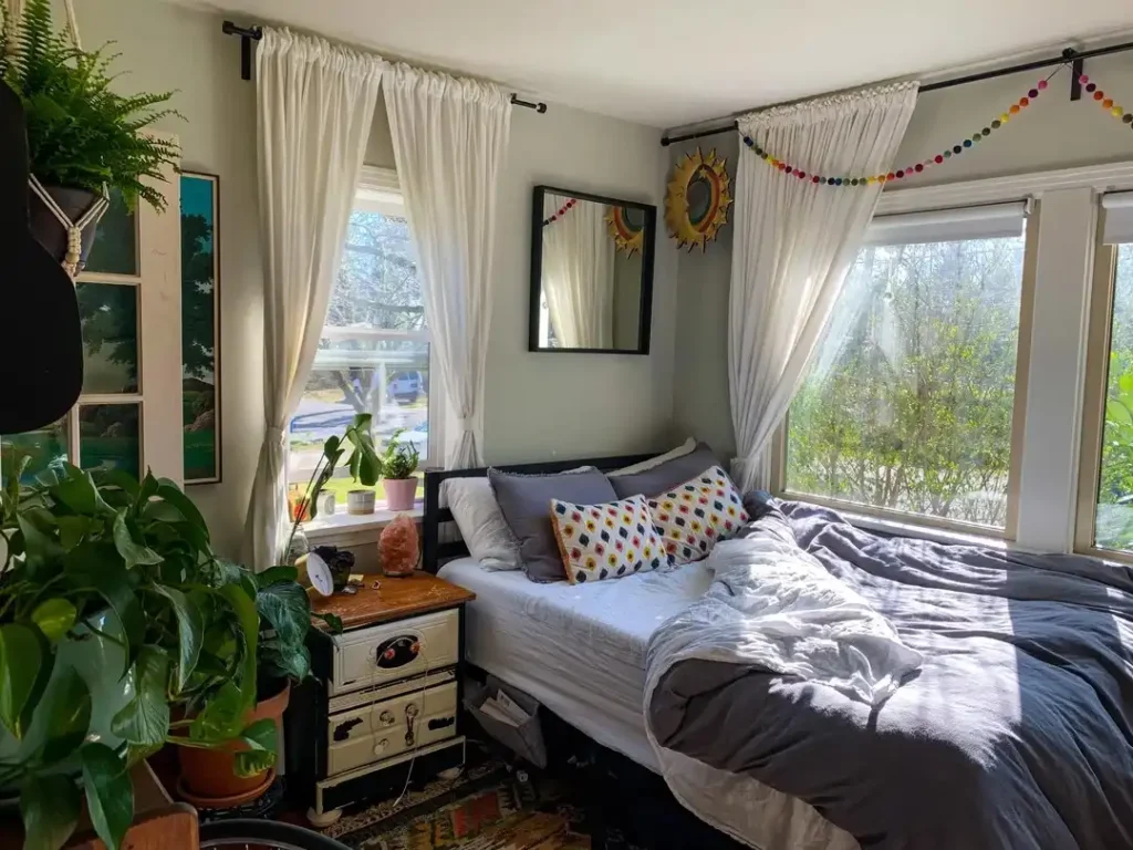 Bright, cozy bedroom with large windows letting in morning sunlight, sheer white curtains, a neatly made bed with gray bedding and patterned pillows, indoor plants, and colorful wall decor creating a warm, airy feel.