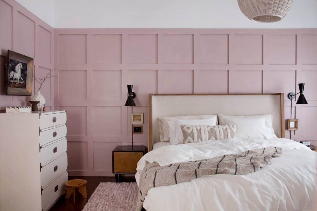 Soft blush pink board-and-batten accent wall behind a neutral upholstered bed, styled with white bedding, black wall sconces, a woven pendant light, and a light wood nightstand for a soft, modern bedroom look.