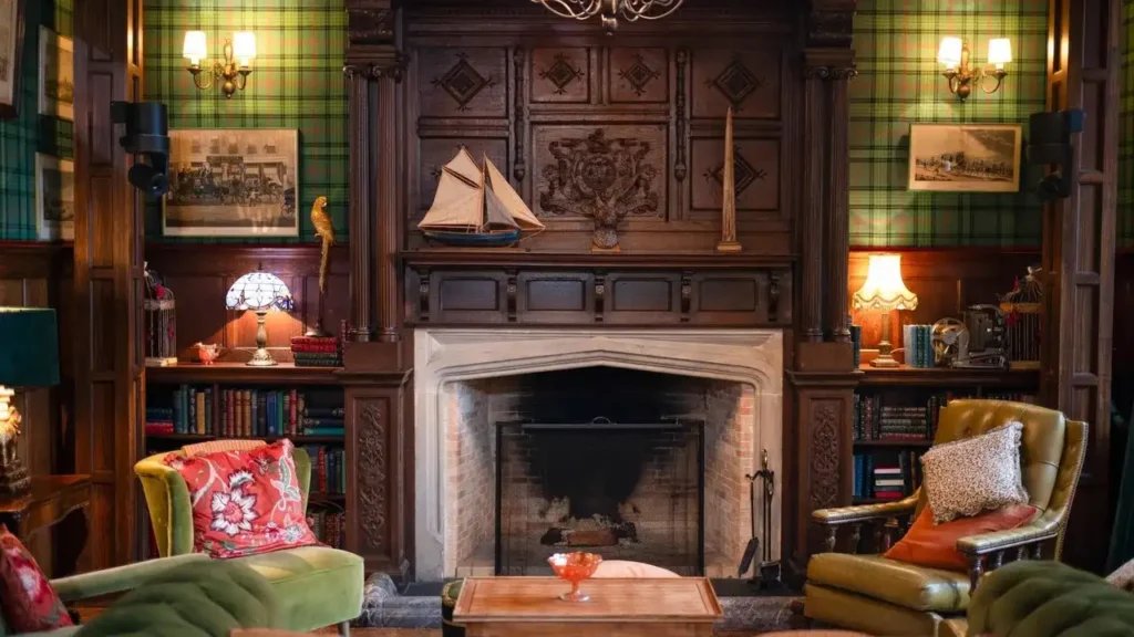 A cozy, Scottish-inspired living room with green tartan wallpaper, dark carved wood paneling, and a large stone fireplace at the center. Velvet armchairs, layered bookshelves, warm table lamps, and eclectic decor create a rich, stately Highlands feel.