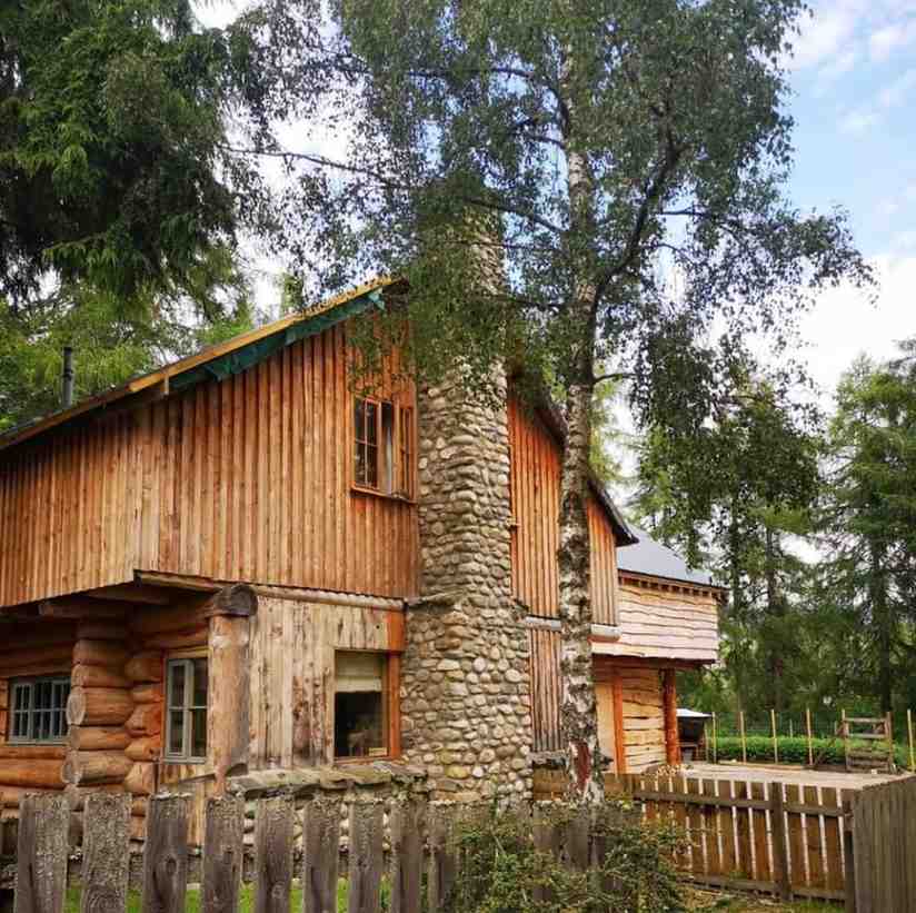 Exterior of Imrich Lodge, a hand-built log home in the Scottish Highlands, featuring natural timber walls, a stone chimney, and mature trees surrounding the house.