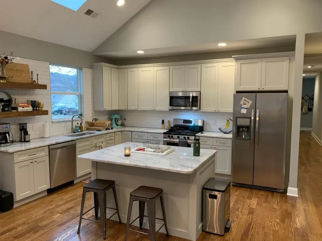 L-shaped kitchen with white cabinets, marble island with seating, stainless steel appliances, subway tile backsplash, and warm wood flooring.