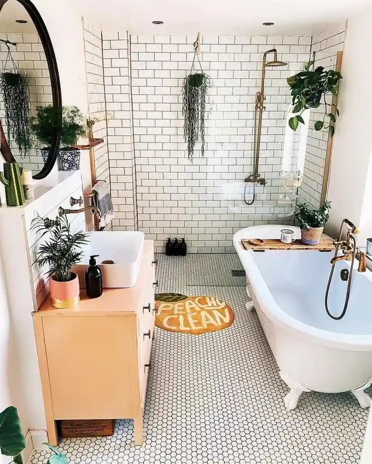 Boho-style bathroom with white subway tile walls, clawfoot tub, brass fixtures, hanging plants, round black mirror, wood vanity, and hex tile floor featuring a “Peachy Clean” rug.