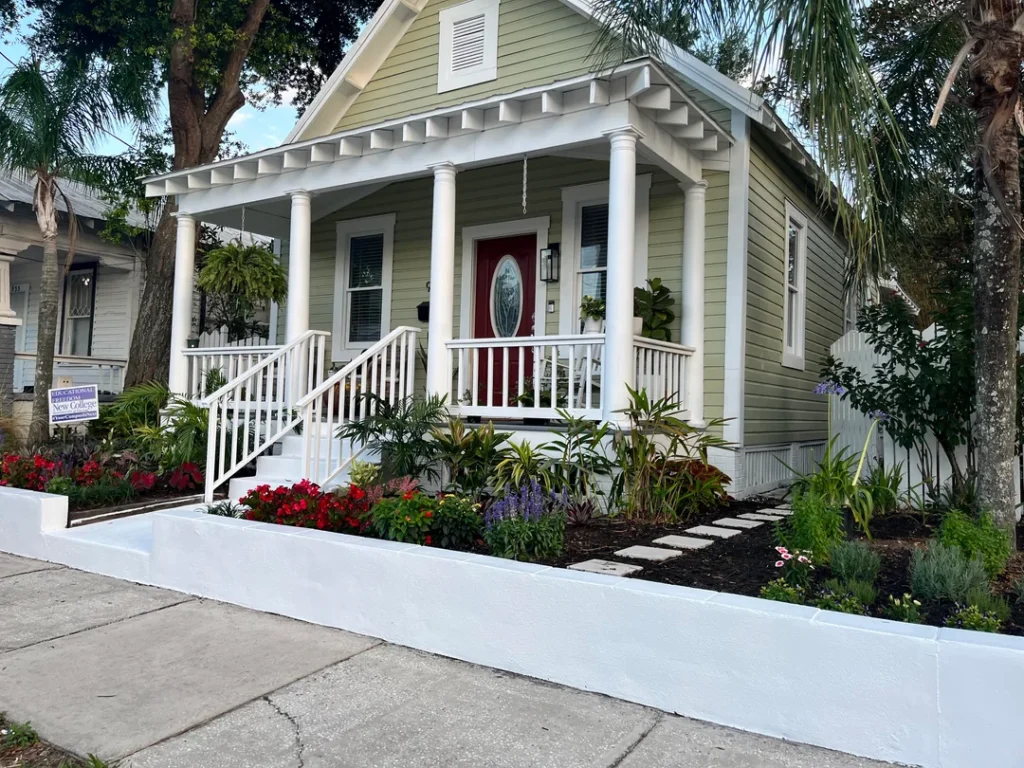 Charming cottage-style home with sage green siding, white porch columns, and a red front door, framed by a raised front yard garden bed with colorful flowers, tropical plants, and a stepping-stone pathway leading to the porch stairs.