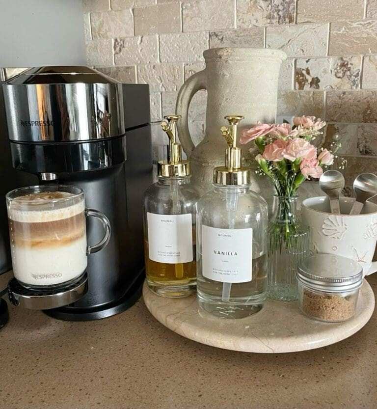 Compact kitchen counter coffee bar featuring a sleek black Nespresso machine brewing a layered latte, glass syrup dispensers with gold pumps, a small vase of pink flowers, and neatly arranged coffee essentials on a round marble tray against a textured tile backsplash.