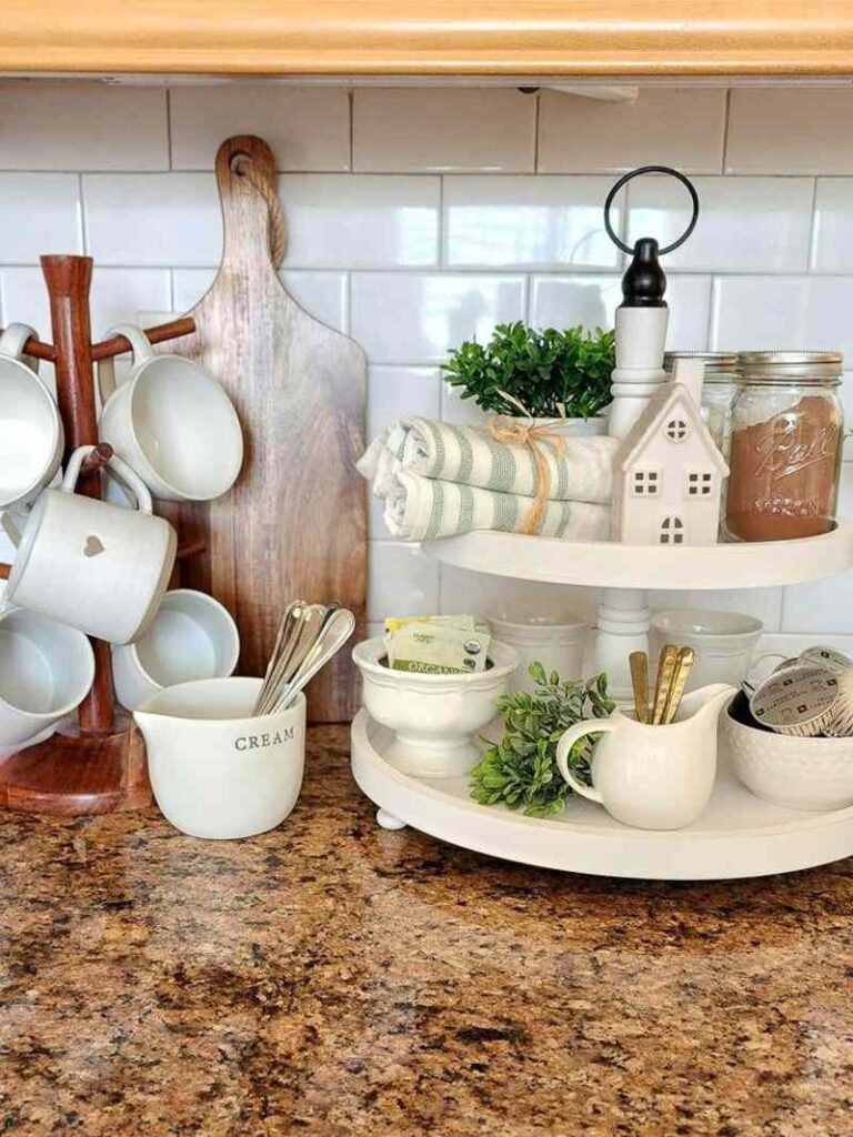 Cozy coffee bar setup with a white two-tier tray holding mugs, creamers, coffee pods, greenery, and farmhouse decor beside a wooden mug stand and cutting board on a warm kitchen countertop.