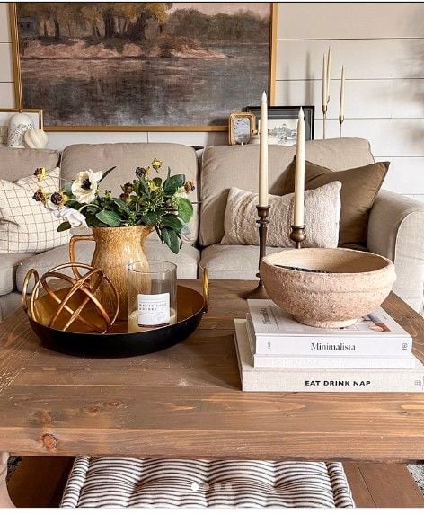 Rustic coffee table centerpiece featuring a textured ceramic vase filled with faux florals, a decorative tray with a candle and gold orb accent, and a large stone bowl styled on stacked coffee table books. Neutral-toned sofas, layered pillows, and warm artwork create a cozy, inviting living room atmosphere.