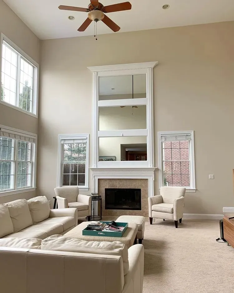 Bright neutral living room with soaring ceilings, beige walls, tall white-trimmed fireplace, large windows, cream sectional sofa, and cozy earth tone decor.