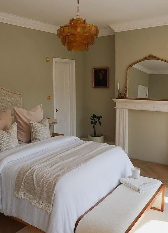 Cozy earth-tone bedroom with soft greige walls, white bedding, blush accent pillows, a cream bench, vintage amber chandelier, and elegant fireplace with an arched mirror.