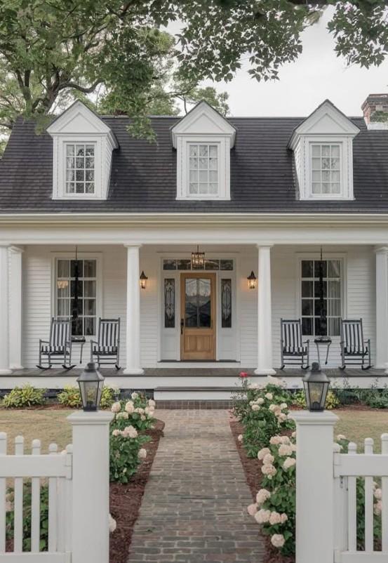 White modern farmhouse exterior with black roof, dormer windows, front porch rocking chairs, brick walkway, and blooming white flowers.