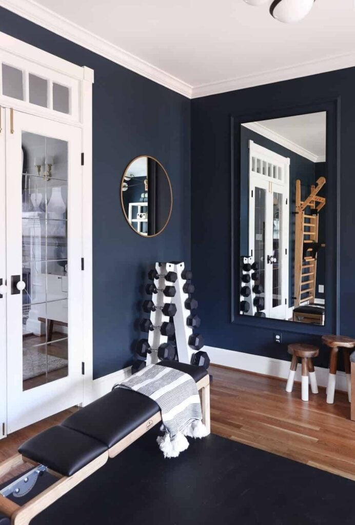 Modern home gym with deep navy blue walls, white trim, hardwood floors, and workout equipment including dumbbells, a bench, mirrors, and wooden fitness bars for a stylish and moody workout space.