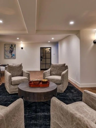 Modern basement sitting area with cream upholstered swivel chairs around a round dark wood coffee table, black area rug, recessed lighting, and soft warm beige walls.