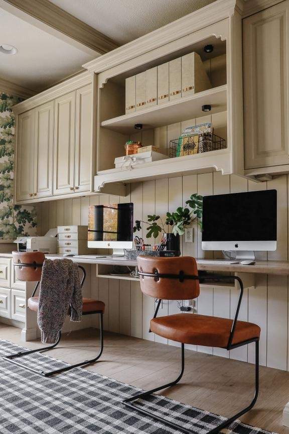 Warm farmhouse-style home office with beige built-in cabinets, wood countertops, leather accent chairs, shiplap walls, and dual desktop computers creating a cozy and organized workspace.
