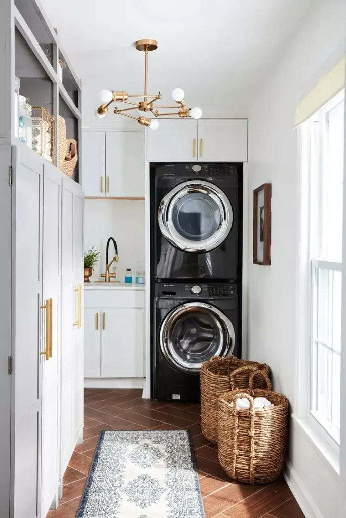 Bright narrow laundry room with stacked black washer and dryer, white cabinets with gold hardware, woven baskets, warm wood herringbone flooring, and a modern brass chandelier.