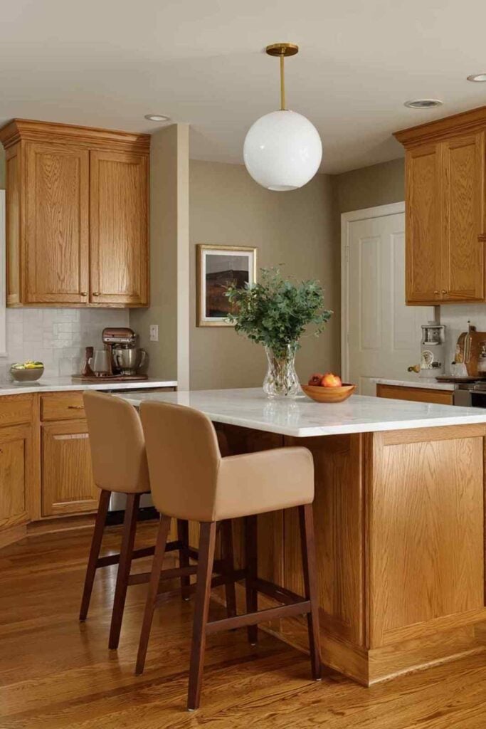 Warm honey oak kitchen cabinets paired with soft greige walls, white quartz countertops, and a cozy kitchen island with tan bar stools and brass pendant lighting.