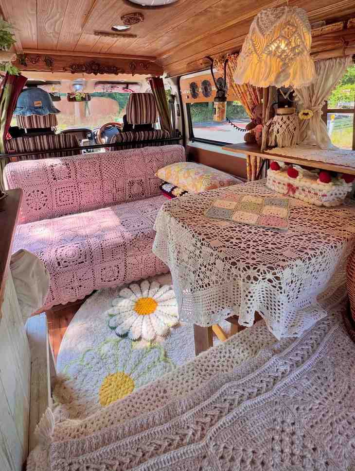 Boho hippie camper interior with cozy crochet blankets covering the sofa, table, and seating, a pink crocheted couch, vintage macramé lamp, daisy rug, warm wood ceiling, colorful curtains, and handmade decor creating a bright, free-spirited van life space.