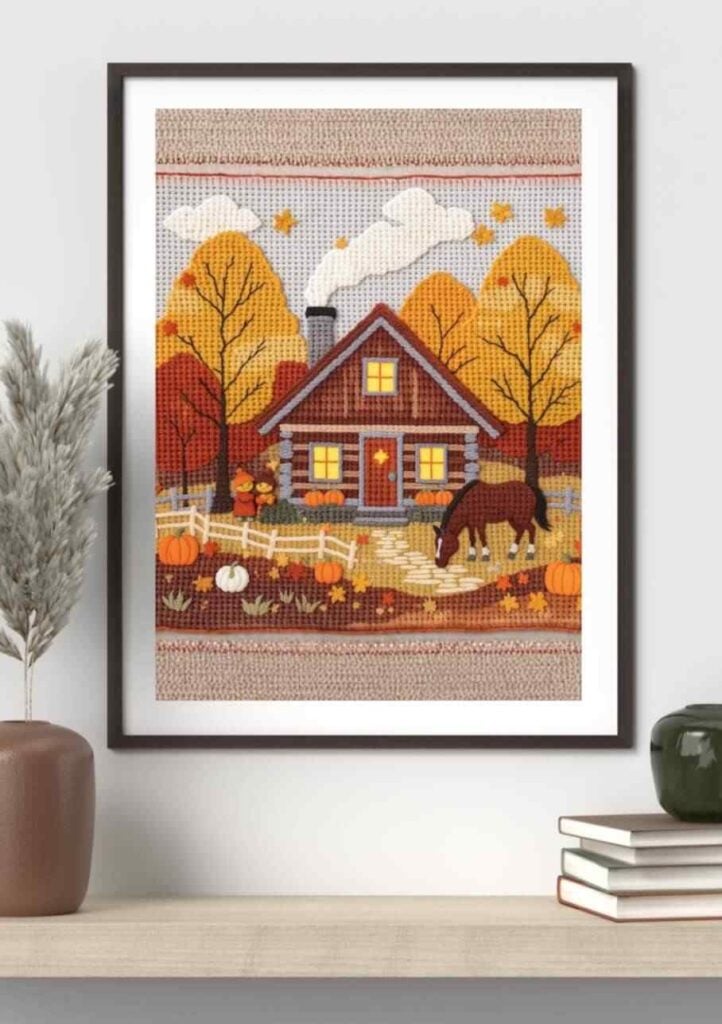 Framed crochet wall art featuring a cozy autumn cabin scene, surrounded by warm-toned trees, pumpkins, and a grazing horse. Textured yarn details create depth, while soft oranges, browns, and creams add a rustic, seasonal charm to this handmade decorative piece.