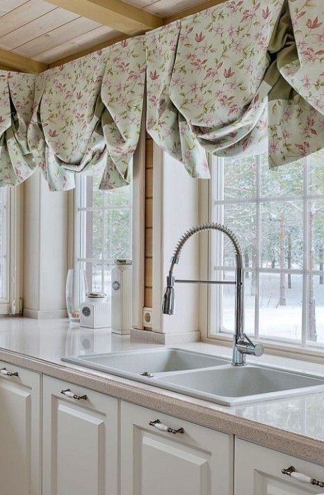 White farmhouse kitchen window dressed with soft floral balloon valances in muted green and pink. The gathered fabric adds cottage charm above large windows, filtering natural light over a white double sink, light countertops, and cream cabinetry. Warm wood ceiling beams complete the cozy, country-inspired kitchen design.