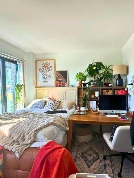 Cozy studio apartment corner featuring a low bed with textured beige bedding, a wooden desk with computer setup, indoor plants, and warm decor accents. Natural light streams through a window beside the bed, while a patterned rug and soft furnishings create a comfortable, stylish small-space living and workspace combo.