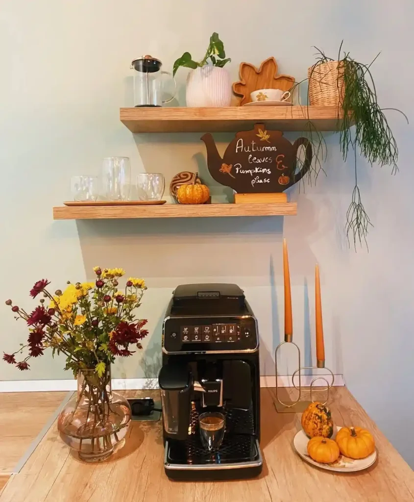 Cozy fall coffee station featuring a black espresso machine on a wooden countertop, styled with fresh autumn flowers, decorative pumpkins, warm candle holders, and floating wood shelves displaying mugs, glassware, greenery, and seasonal fall accents.