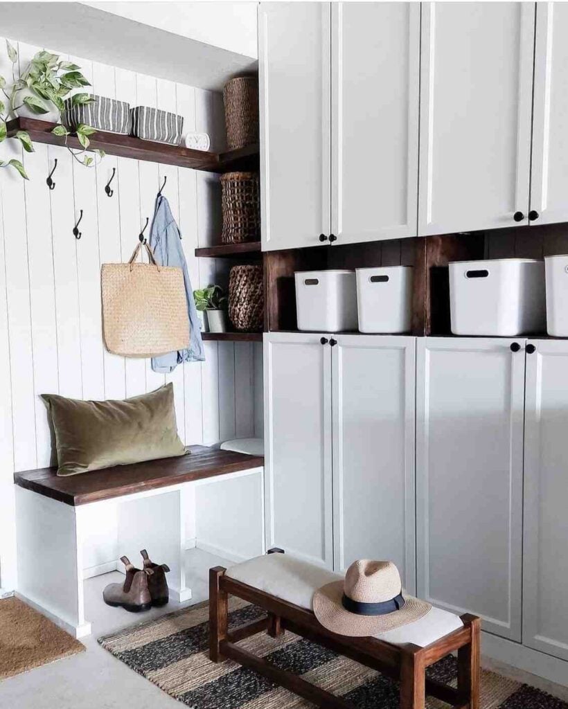 Bright mudroom with white built-in cabinets, dark wood benches and shelves, wall hooks, woven baskets, storage bins, greenery, and a striped rug.