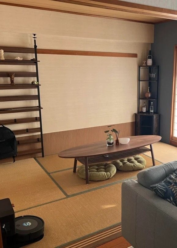 Minimal tatami living room with a low wooden table and round floor cushions, open wall shelving on one side, and a small glass display cabinet. Warm wood tones, clean lines, and natural light create a calm, uncluttered Japanese-style space.