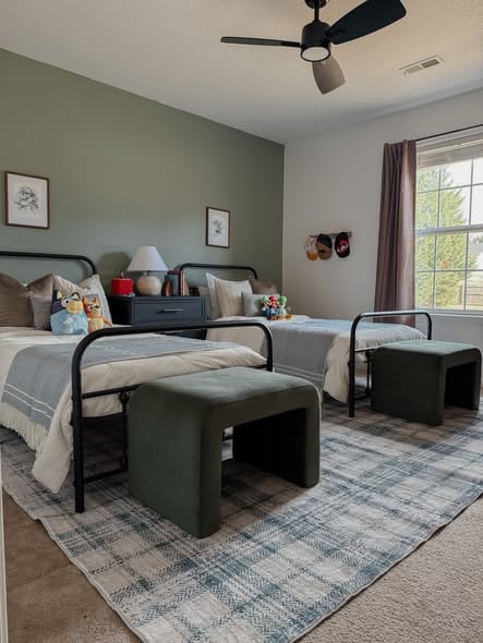 Two boys’ bedroom with twin black metal beds, sage green accent wall, gray plaid rug, storage benches, and soft neutral bedding.