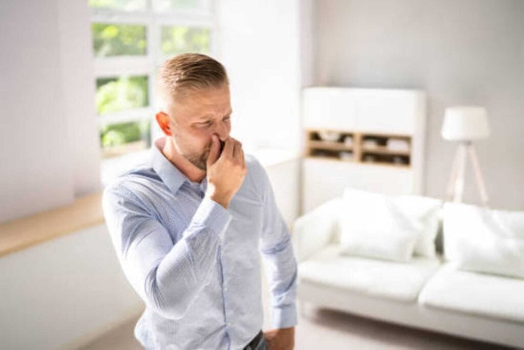 Man pinching his nose in disgust while standing in a bright, neatly furnished living room.