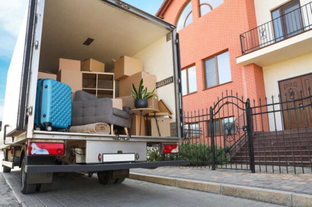 Moving truck loaded with boxes, furniture, a blue suitcase, and a houseplant parked outside a brick home.