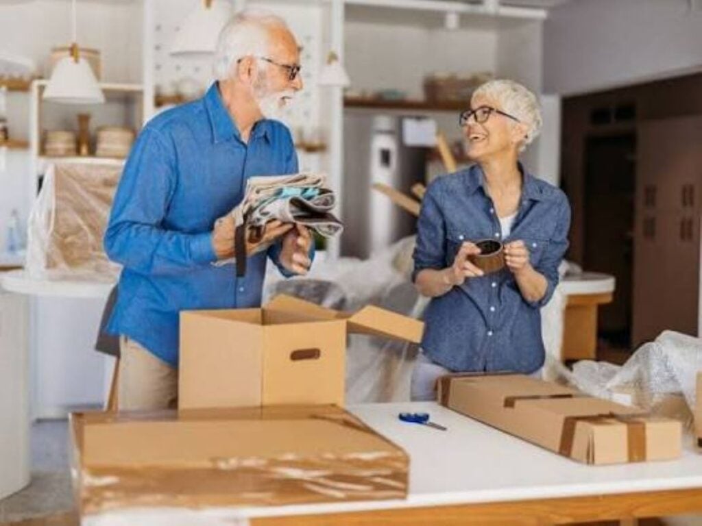 Older couple smiling while decluttering their home, with cardboard boxes and a stack of magazines on the table.