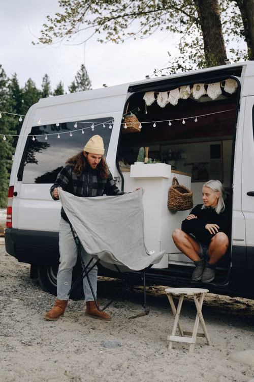 A couple beside a white camper van in a wooded area, with one person unfolding a camping chair while the other sits in the open doorway.