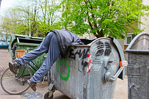 A person leaning headfirst into a large outdoor dumpster while searching through its contents, with a bicycle parked nearby.