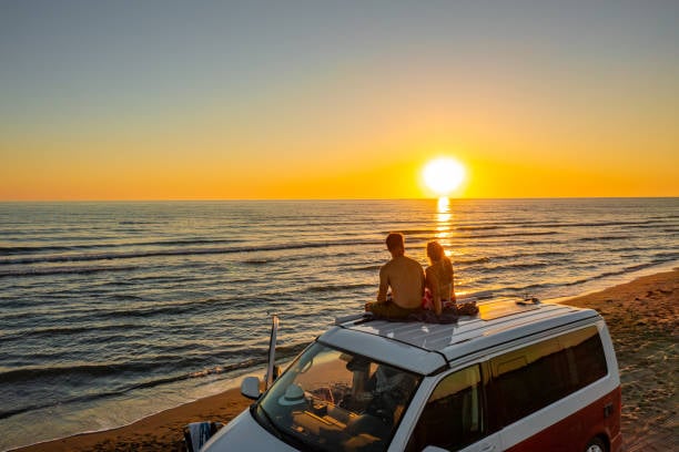 Two people sitting on the roof of a camper van parked beside the beach, watching the sun set over the ocean.