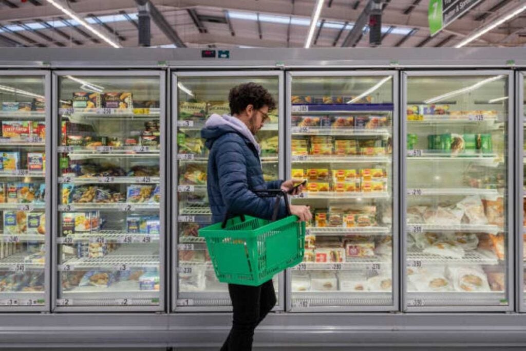 A man carrying a green shopping basket checks his phone while standing in front of frozen food cases at a grocery store.
