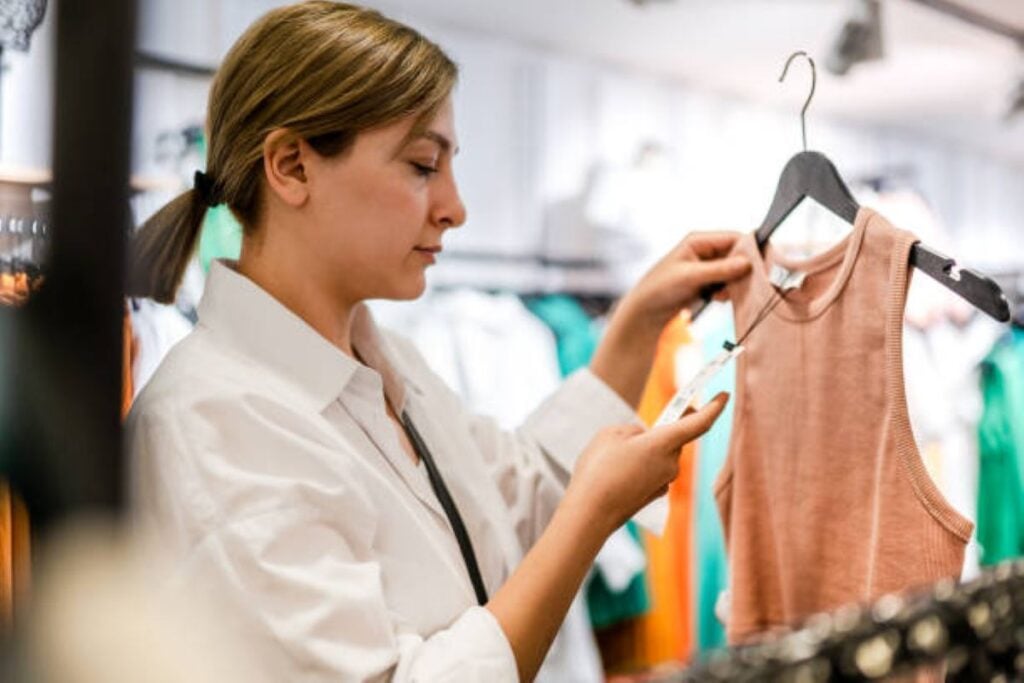 Woman shopping in a clothing store, holding a peach sleeveless top on a hanger while checking its price tag.