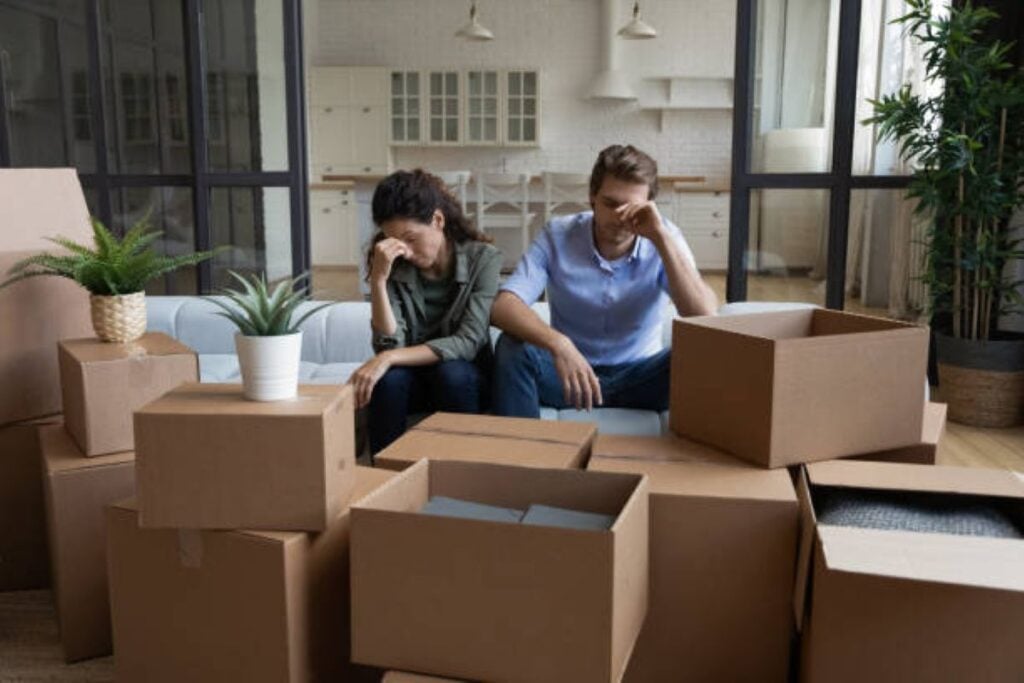 Two people sit on a couch looking stressed and tired, surrounded by packed and open cardboard moving boxes in a bright living room.