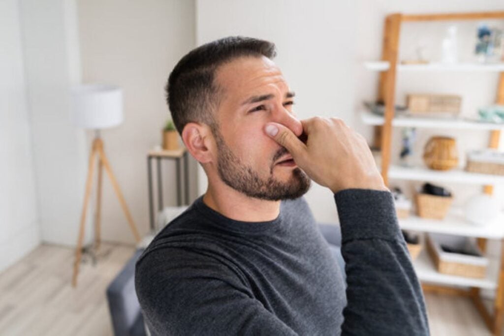 A man sitting in a living room pinches his nose with a disgusted expression, reacting to an unpleasant smell in his home.