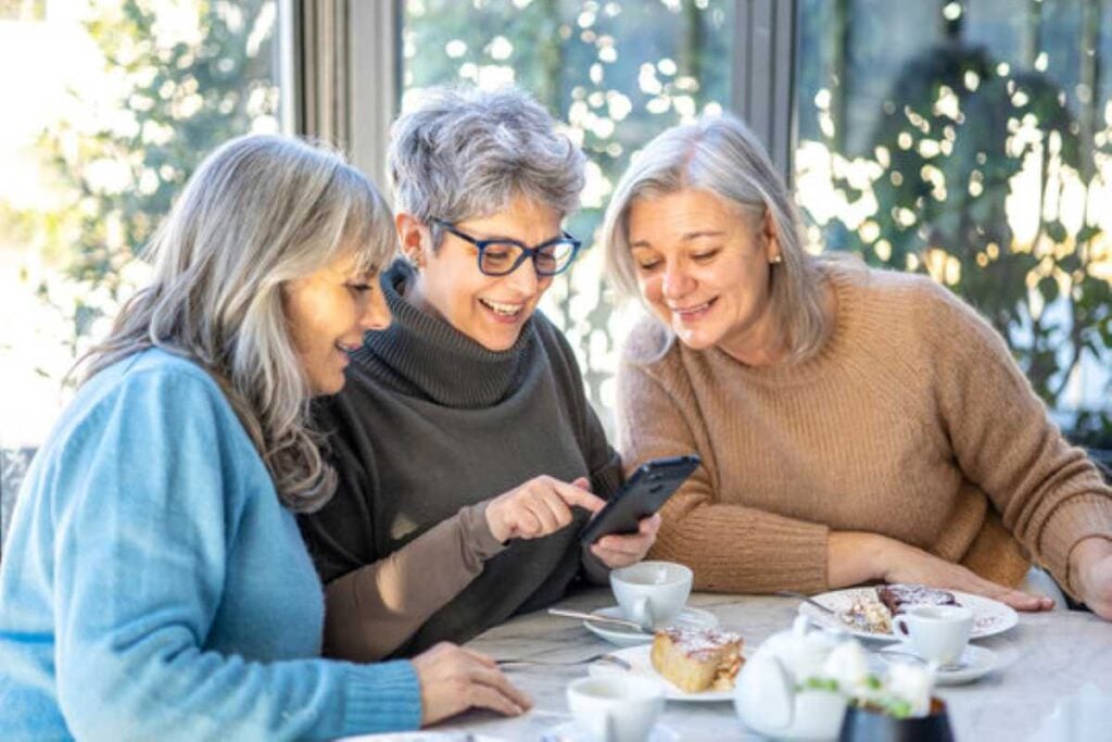 Three older women laughing together over a smartphone while enjoying coffee and cake at a table.