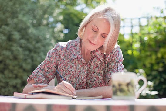 Older woman sitting outdoors and writing in a journal at a table surrounded by greenery.
