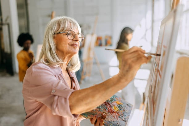 Older woman painting on a canvas in a bright art studio while holding a painter’s palette.