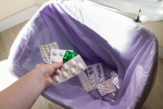 Hand dropping blister packs of unused medication into a trash bin lined with a purple bag.